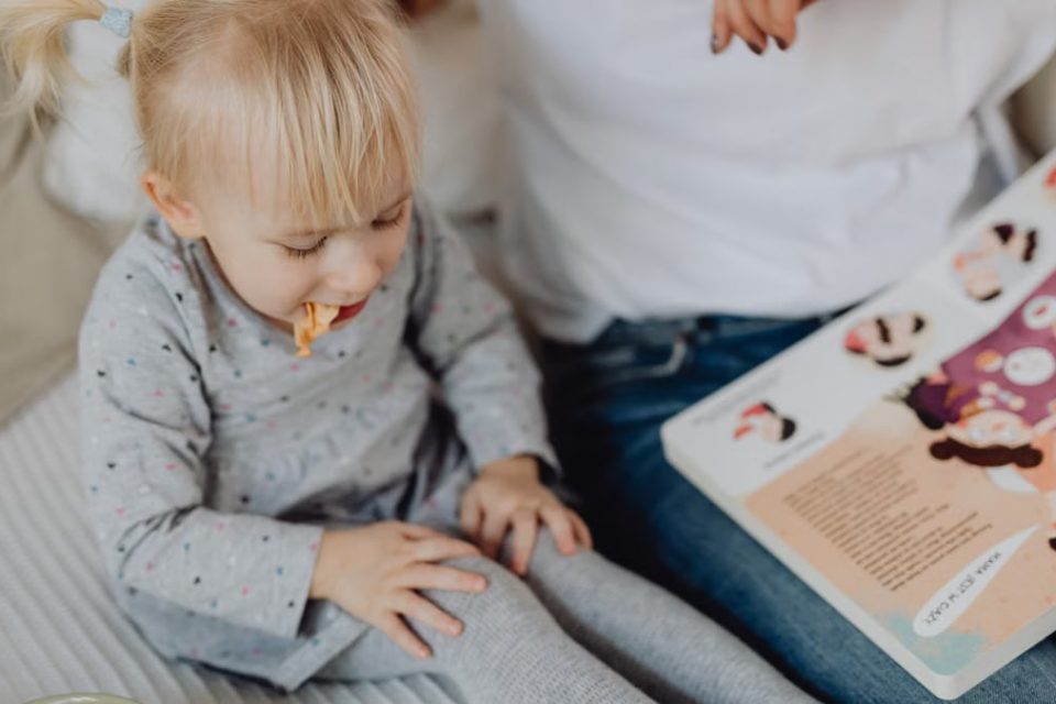 Cute toddler with ponytails enjoying a snack next to a book and parent. Cozy indoor scene.