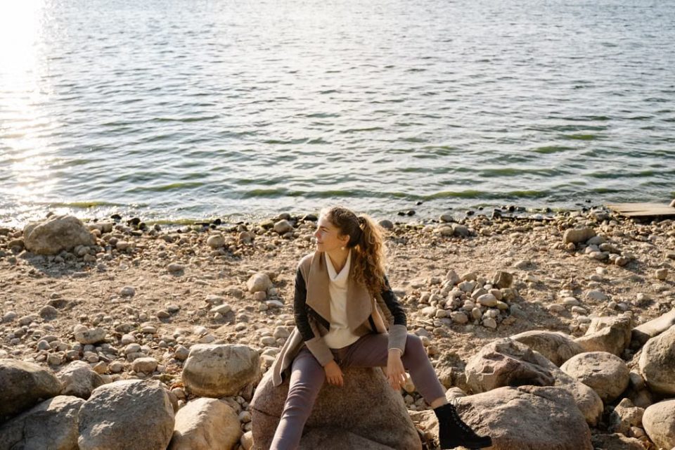 A woman sits on a rocky shoreline, gazing at the serene lake view during a sunny day.